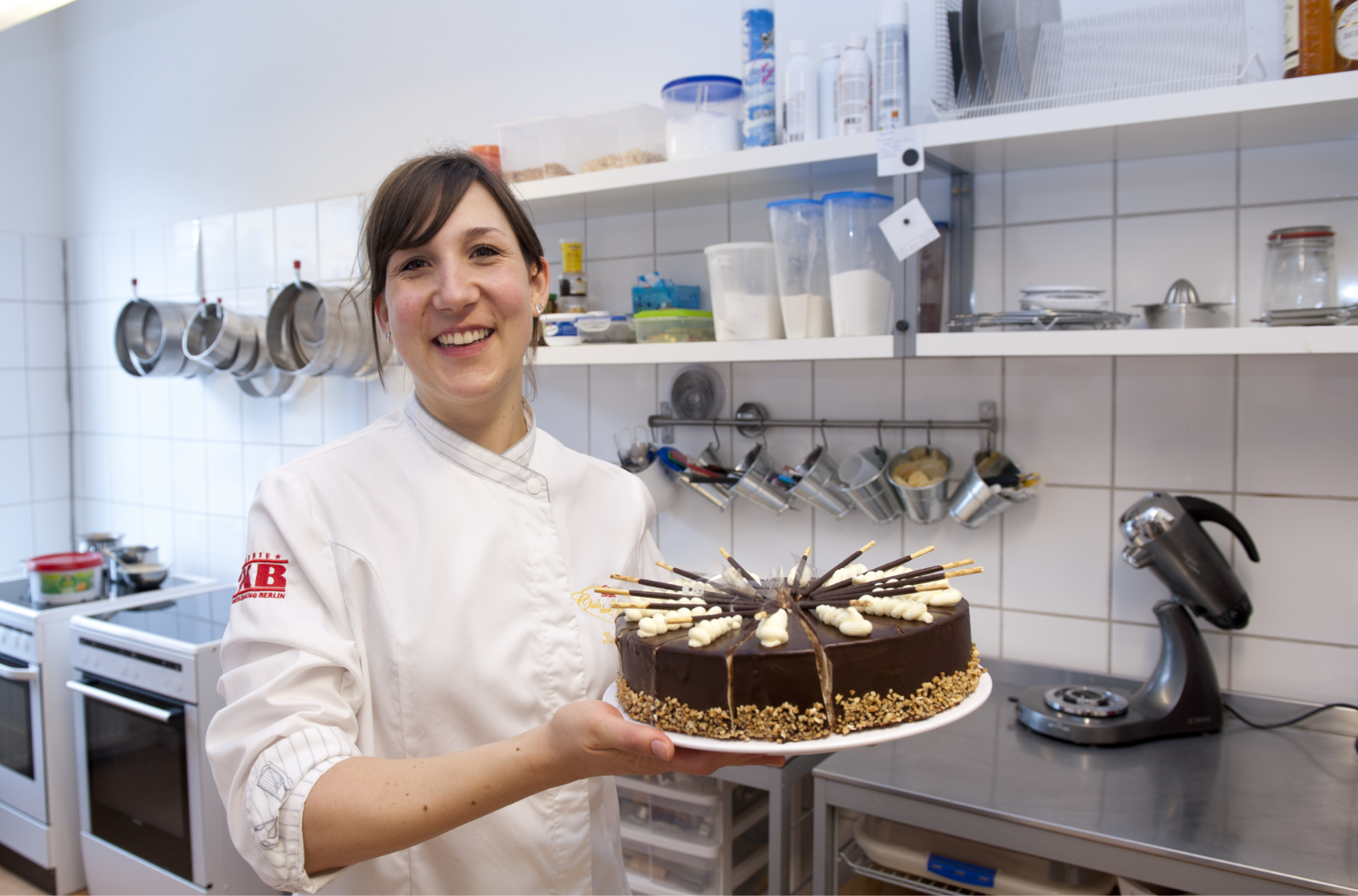 Ein Kochhemd tragende junge Frau in einer Großküche präsentiert lächelnd eine Torte.